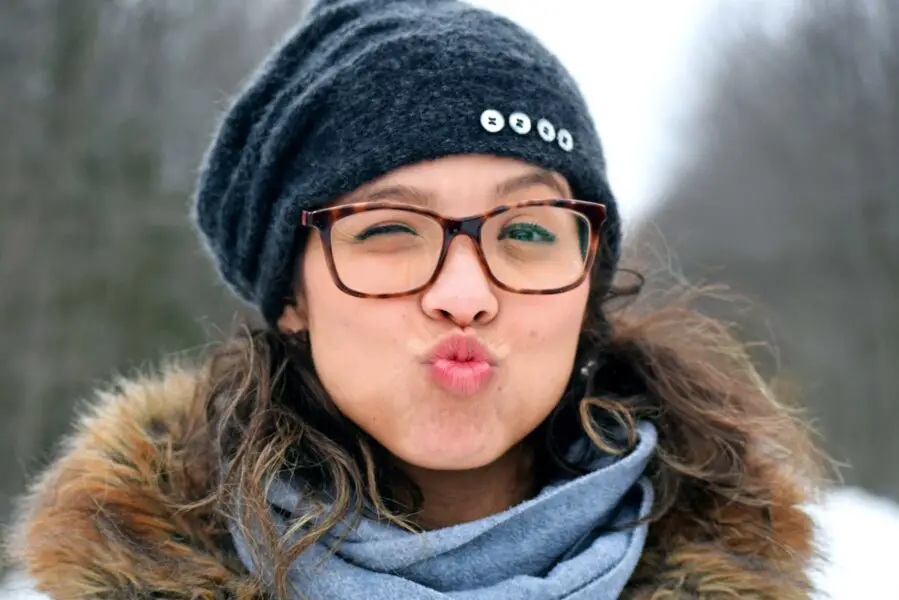 A woman in glasses wearing winter clothes puckers her lips and winks.