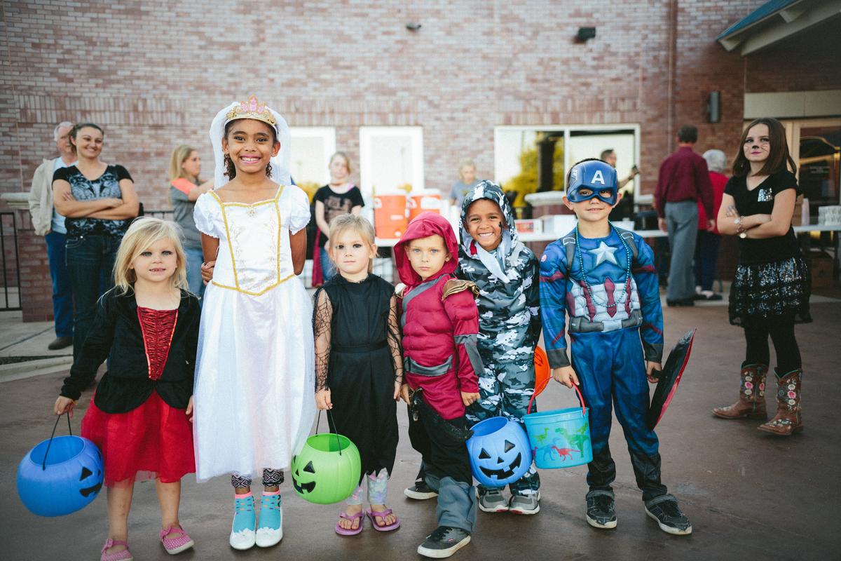 The image shows a group of children dressed in Halloween costumes standing in front of a table filled with festive snacks and treats, including apple slice monster mouths, mummy dogs, pumpkin-shaped cheese balls, candy corn, and Mexican candies.